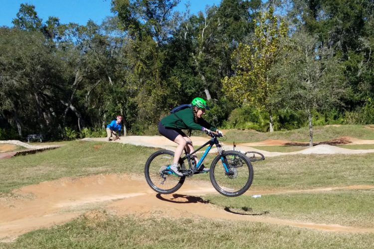 A young person wearing a green helmet and a dark green sweater is mid-air on a mountain bike, performing a jump over a dirt track in a park. In the background, another individual is visible, observing the scene. The area is surrounded by greenery, with trees and open grass patches.