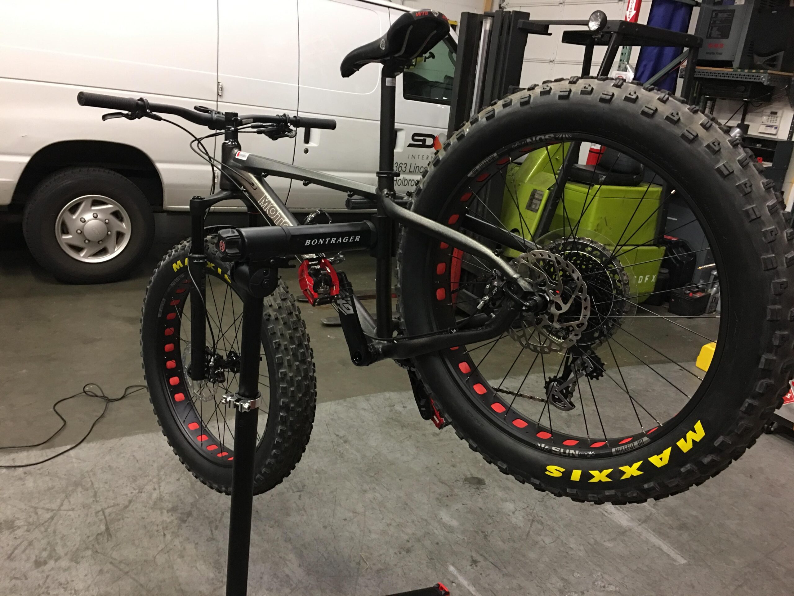 Motobecane NightTrain Bullet: A fat tire mountain bike is positioned on a repair stand in a garage workspace. The bike features wide tires with red accents, a sleek frame, and is equipped with disc brakes. In the background, there is a white van and various tools and equipment typical of a bike shop setting.