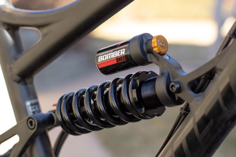 Close-up view of a motorcycle shock absorber featuring a black coil spring and a cylindrical reservoir with branding. The shock is mounted on a black frame, showcasing its components and design details.