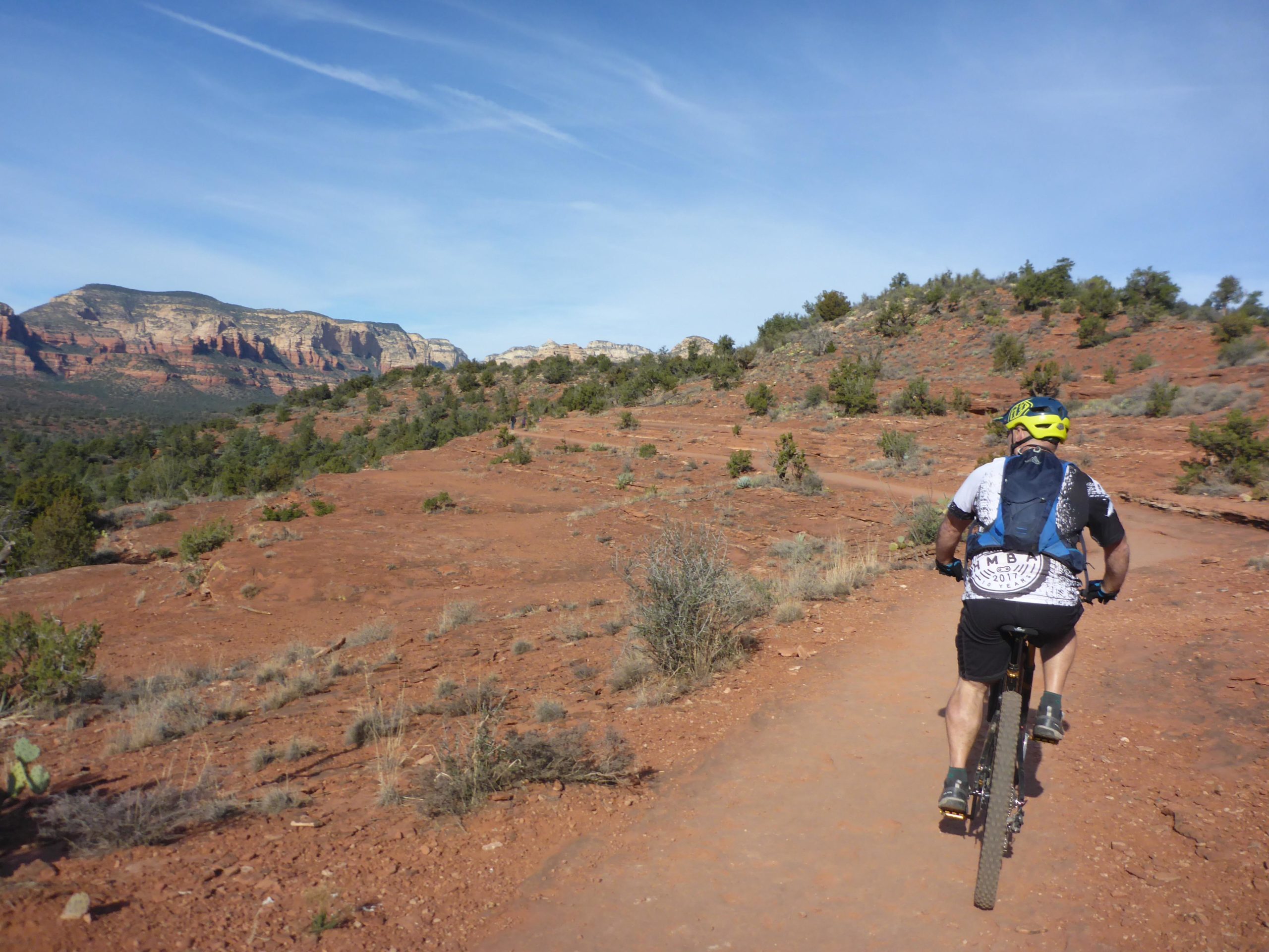 A person riding a mountain bike on a dirt trail, surrounded by red rocky terrain and green shrubs. The background features rocky cliffs and a blue sky with wispy clouds. Chuck Wagon mountain bike trail.