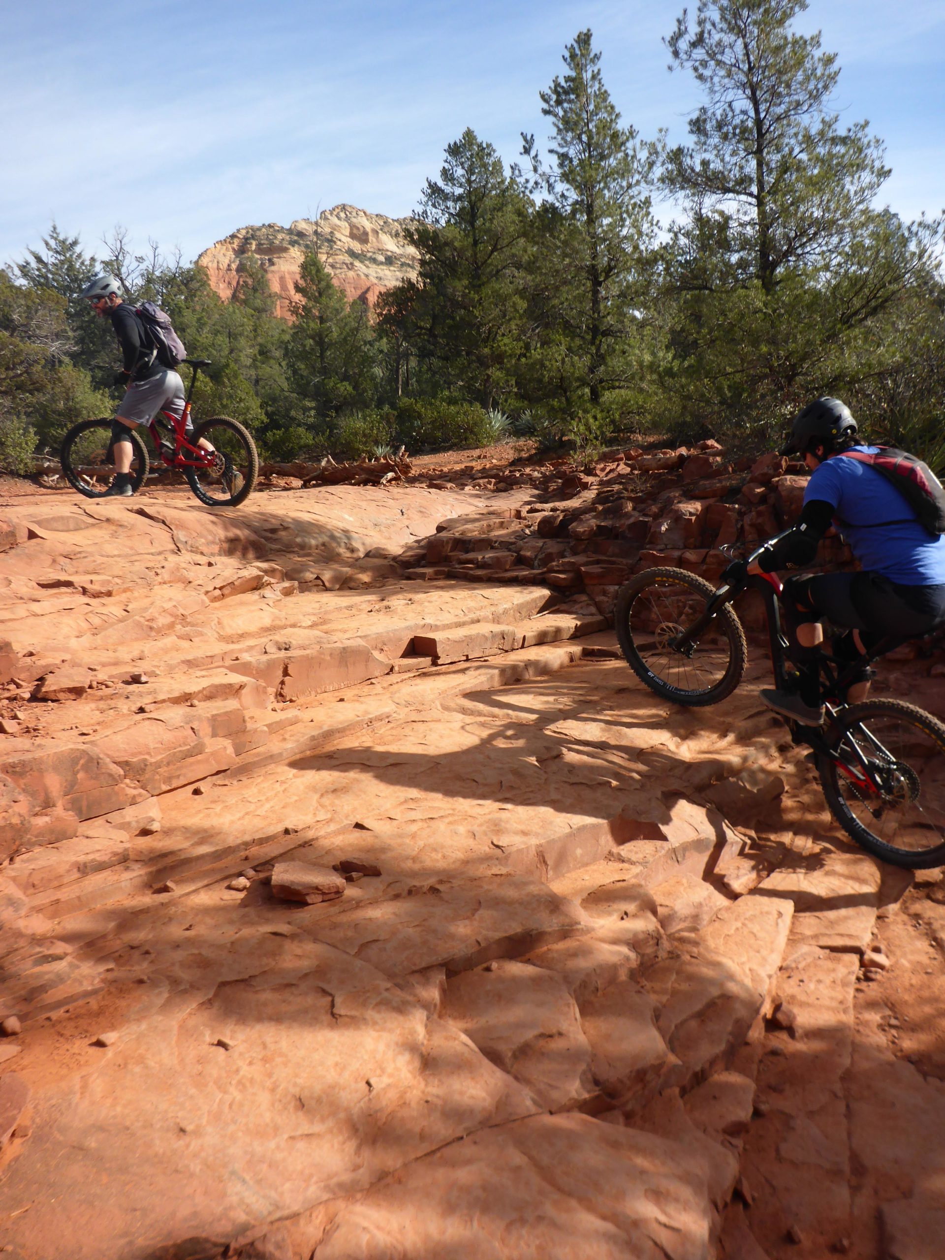 Two mountain bikers navigate a rocky trail surrounded by trees and red rock formations. The cyclist in the foreground wears a blue shirt and helmet, while the rider in the background is dressed in gray and has a backpack. The landscape features rugged terrain under a clear blue sky. Chuck Wagon mountain bike trail.