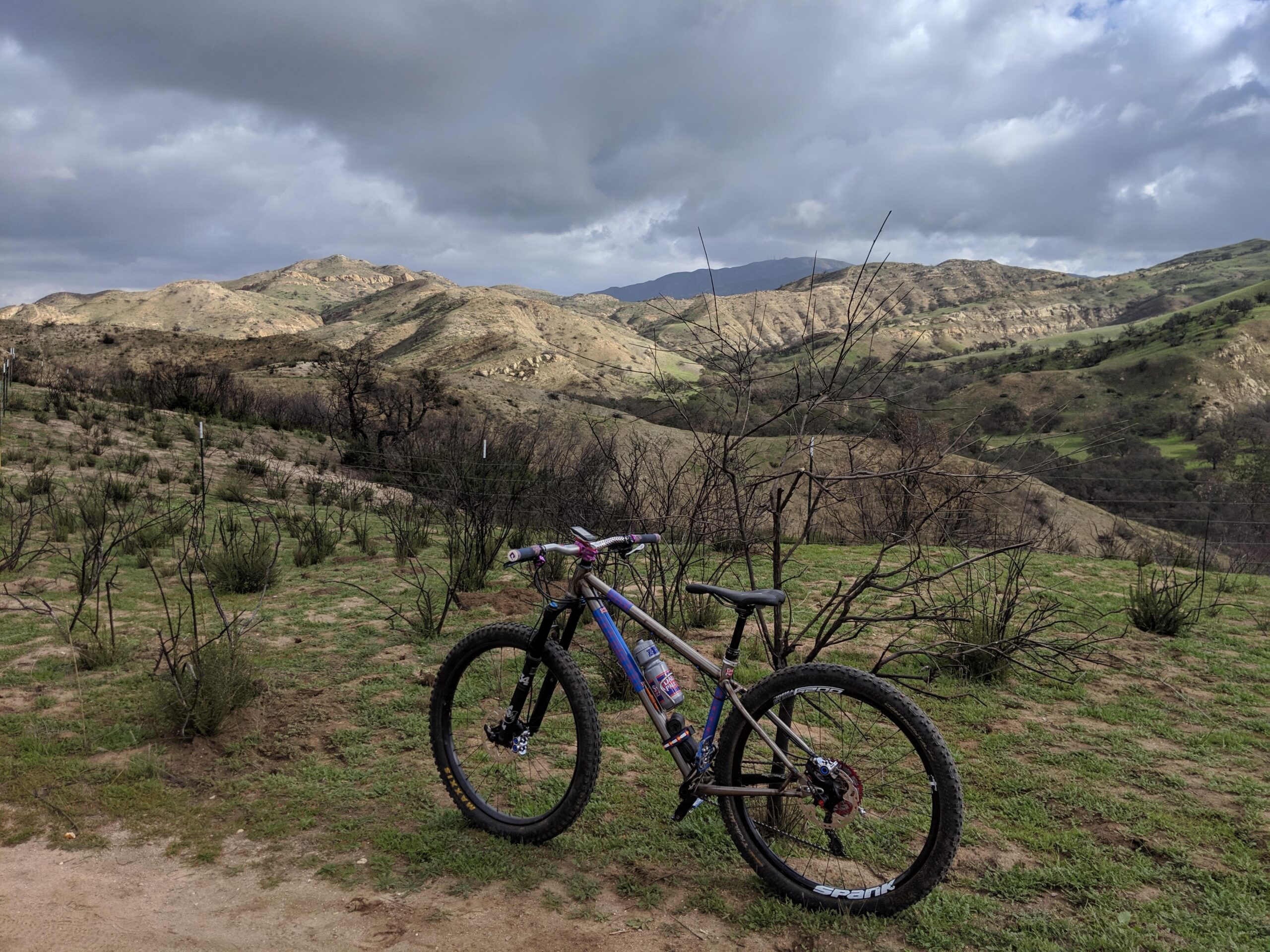 Reeb REEBdikeylous: A mountain bike resting on a dirt trail with a backdrop of rolling hills and a cloudy sky. The landscape features sparse vegetation, some areas of burnt foliage, and distant mountains.