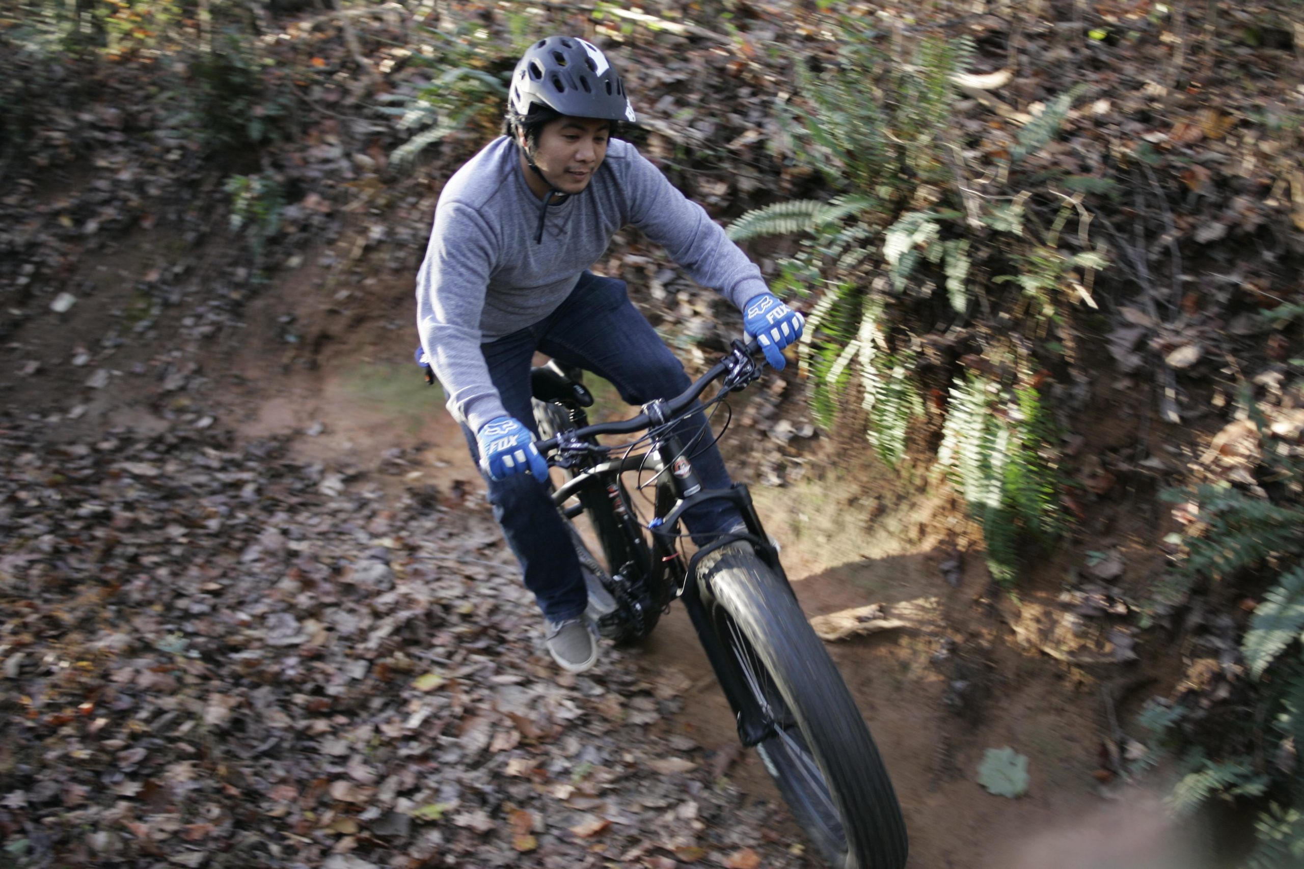 A person wearing a helmet and gloves is riding a mountain bike on a dirt trail surrounded by autumn leaves and greenery. The cyclist is leaning slightly forward as they navigate a curve, showcasing their skill in handling the bike. Hobby Park mountain bike trail.