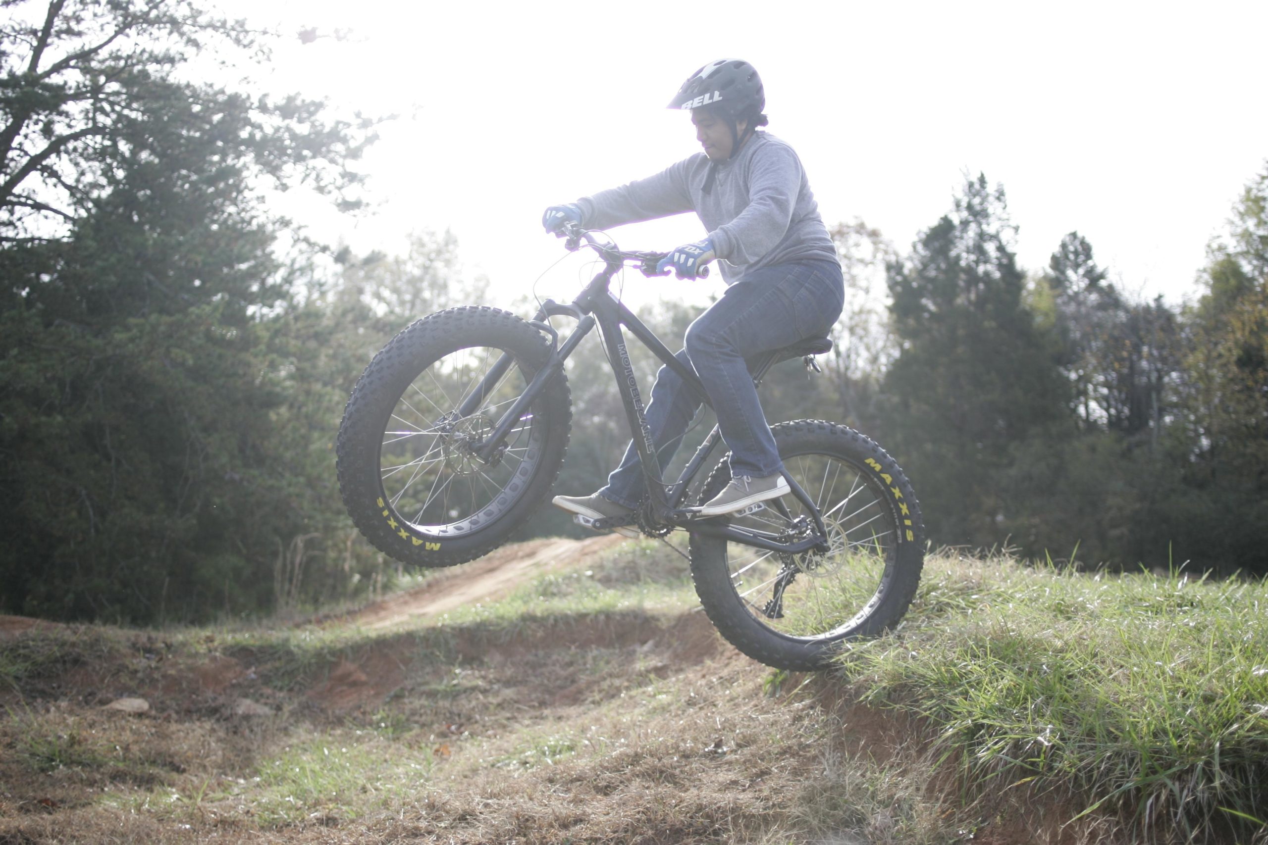 A person in a helmet and long sleeves rides a fat tire bike, performing a wheelie over a dirt ramp in an outdoor setting. The background features trees and grassy terrain, with a bright sky. Hobby Park mountain bike trail.