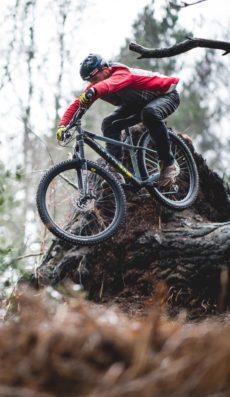 Marin San Quentin 2: A mountain biker in a red jacket and black gear performs a jump over a fallen tree in a wooded area, showcasing an action-packed moment in nature.