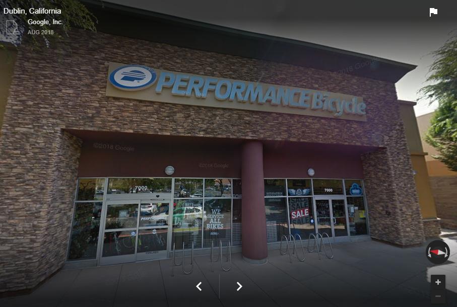 Storefront of Performance Bicycle located in Dublin, California, featuring a stone facade and large sign. The entrance has glass doors showing displayed bicycles and promotional signage, including "WE SERVICE ALL BIKES" and sale announcements. Bike racks are visible in front of the store.