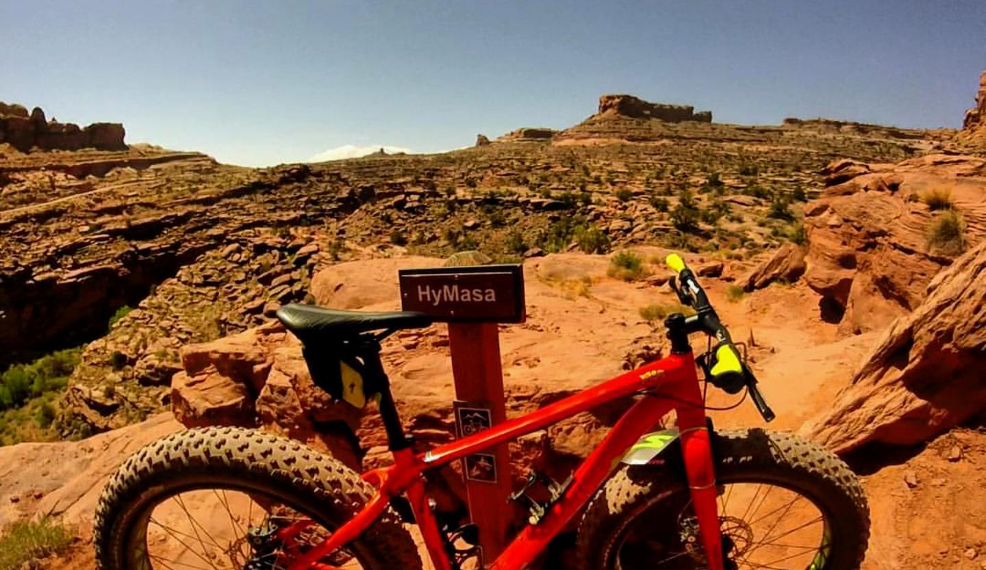 A red mountain bike is parked next to a trail sign that reads "HyMasa," set against a backdrop of rugged, rocky terrain under a clear blue sky. The landscape features layered rock formations and sparse vegetation, typical of a desert environment. Amasa Back Area mountain bike trail.