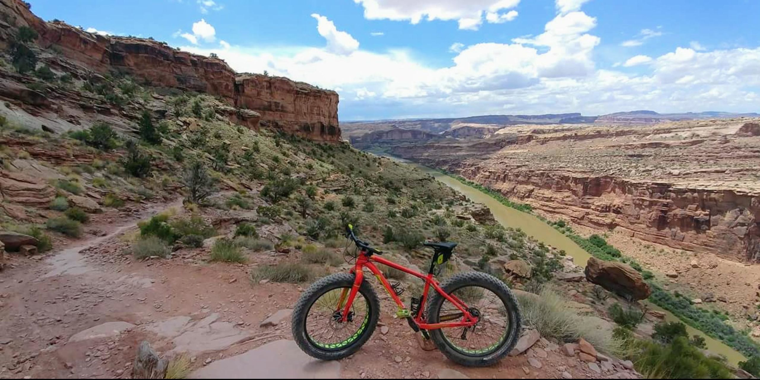 A bright red mountain bike with wide tires is positioned on a rocky trail overlooking a winding river nestled between towering cliffs under a partly cloudy sky. The landscape features shrubs and sparse vegetation, with distant hills and rock formations visible in the background. Porcupine Rim mountain bike trail.