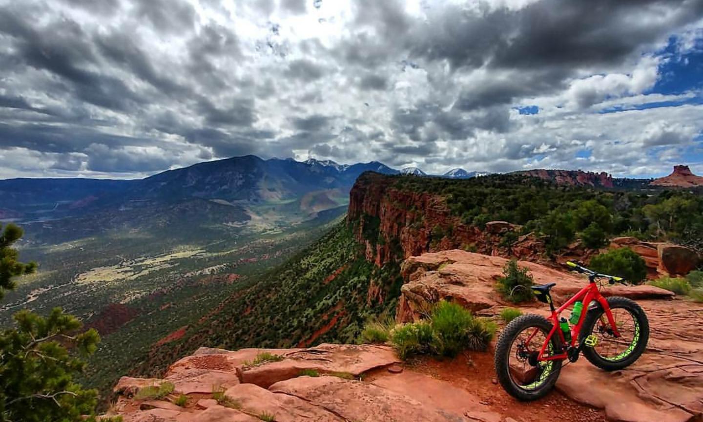 A brightly colored mountain bike resting on a rocky ledge overlooking a vast valley, with rolling hills and distant mountains under a dramatic sky filled with clouds. The landscape features green vegetation and red rock formations, showcasing the beauty of nature. Porcupine Rim mountain bike trail.