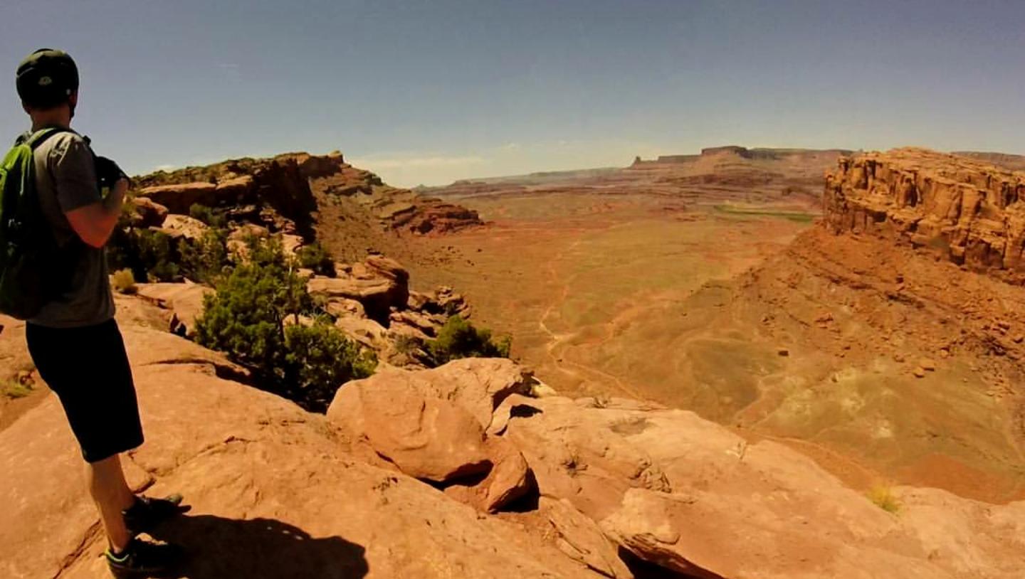 A person standing on a rocky ledge overlooking a vast desert landscape, with red rock formations and a clear blue sky in the background. The scene captures the beauty of nature and the rugged terrain. Amasa Back Area mountain bike trail.