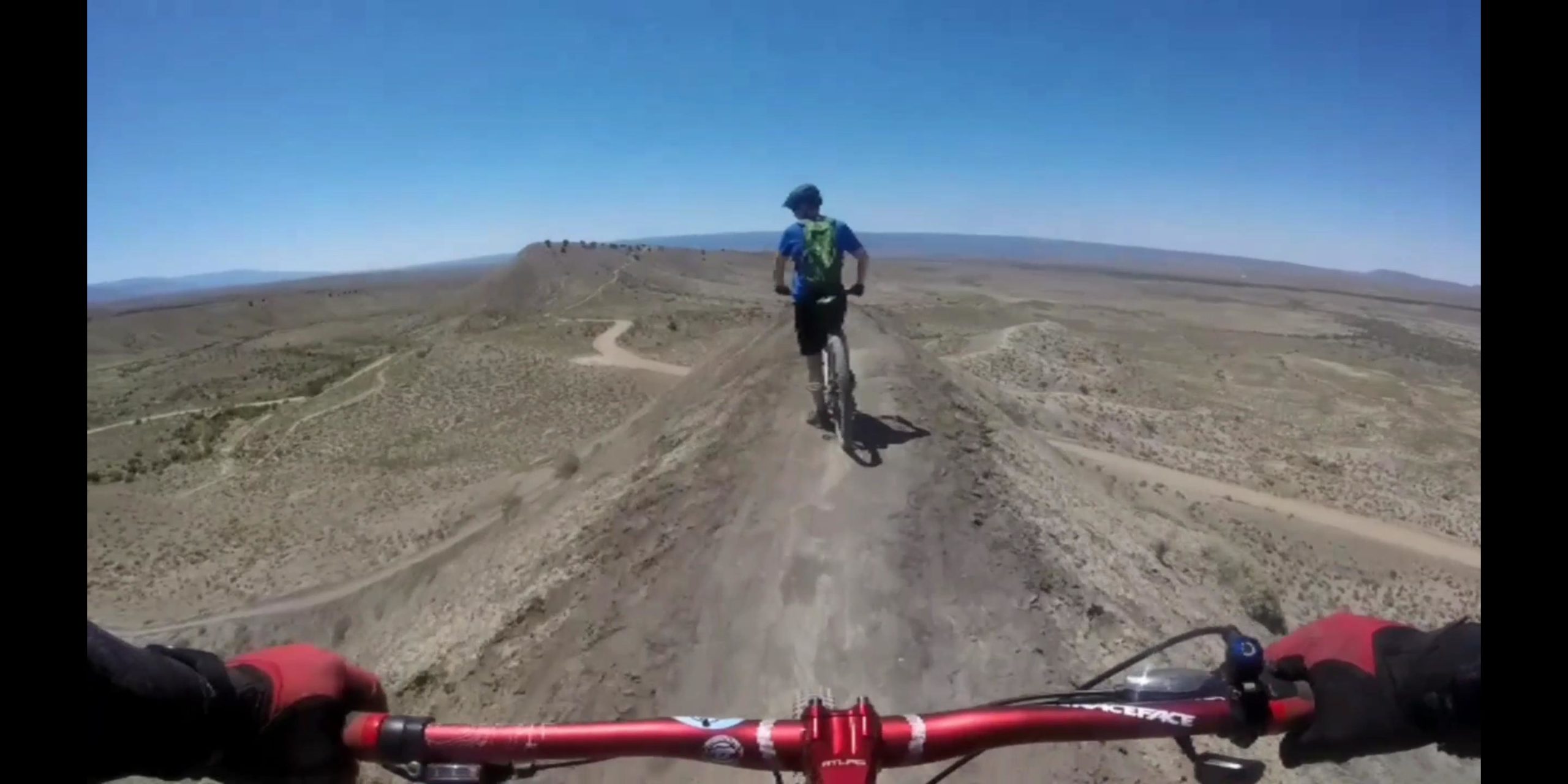 Mountain biker on a rocky trail overlooking a vast desert landscape under a clear blue sky. The scene captures a steep incline, with winding paths visible in the distance, showcasing an adventurous outdoor biking experience. The biker is wearing a helmet and a backpack, indicating preparation for an off-road ride. 18 Road Trails / North Fruita Desert mountain bike trail.