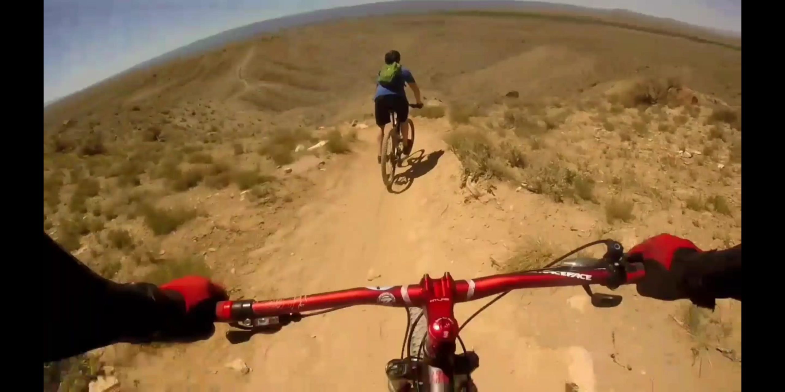 A mountain biker is descending a dirt trail in a rugged landscape. The handlebars of the bike are visible in the foreground, while a second biker can be seen in the distance riding along the trail. The environment is arid with dry dirt and sparse vegetation under a clear blue sky. 18 Road Trails / North Fruita Desert mountain bike trail.
