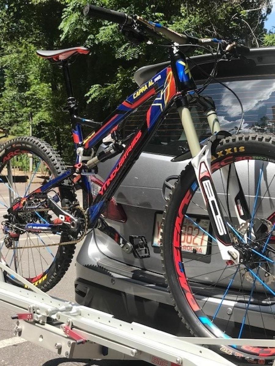YT Capra: A colorful mountain bike is mounted on a bike rack attached to the back of a car, set against a backdrop of green trees. The bike features vibrant red, blue, and black colors, with distinct tire branding. Sunlight casts a bright shine on the bike, highlighting its details.
