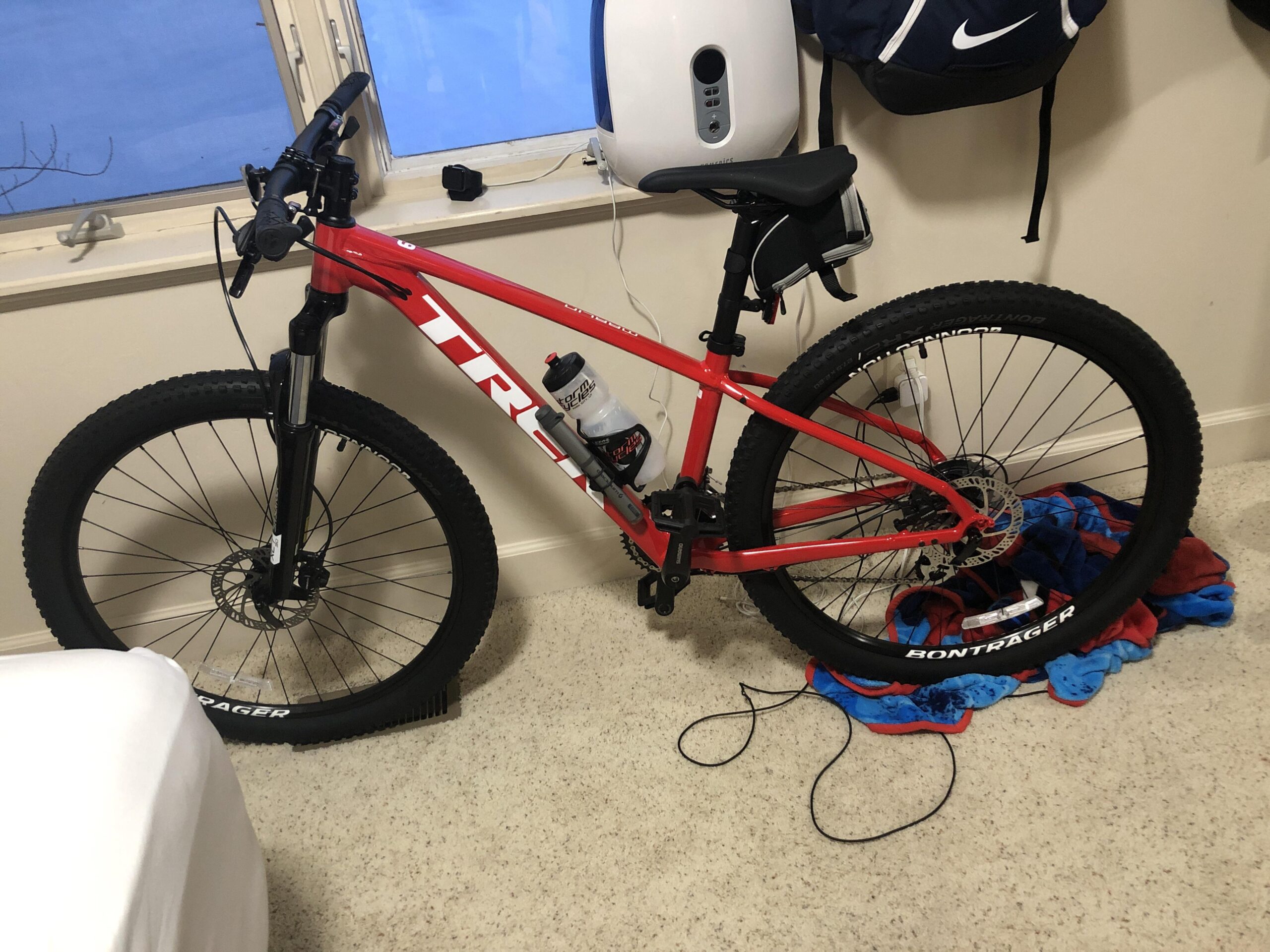 Trek Marlin 6: A red Trek mountain bike leaning against a wall in a room, with a water bottle attached to the frame. The bike is positioned next to a blue and red towel on the floor, and there's a bed with white bedding partially visible in the foreground. A window is visible in the background, reflecting daylight.