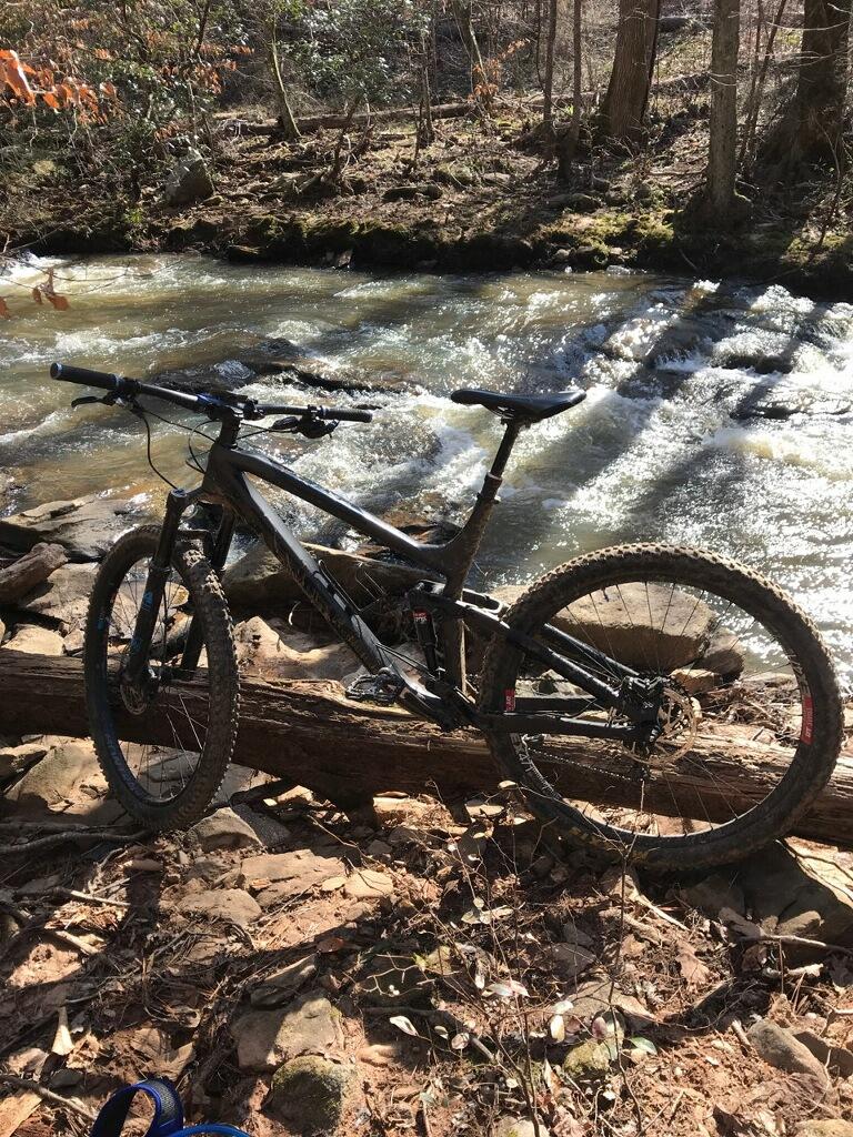 A mountain bike resting against a fallen log by a flowing stream, surrounded by rocks and trees in a natural setting. Sunlight filters through the trees, creating a peaceful outdoor scene. Cochran Mill Park mountain bike trail.