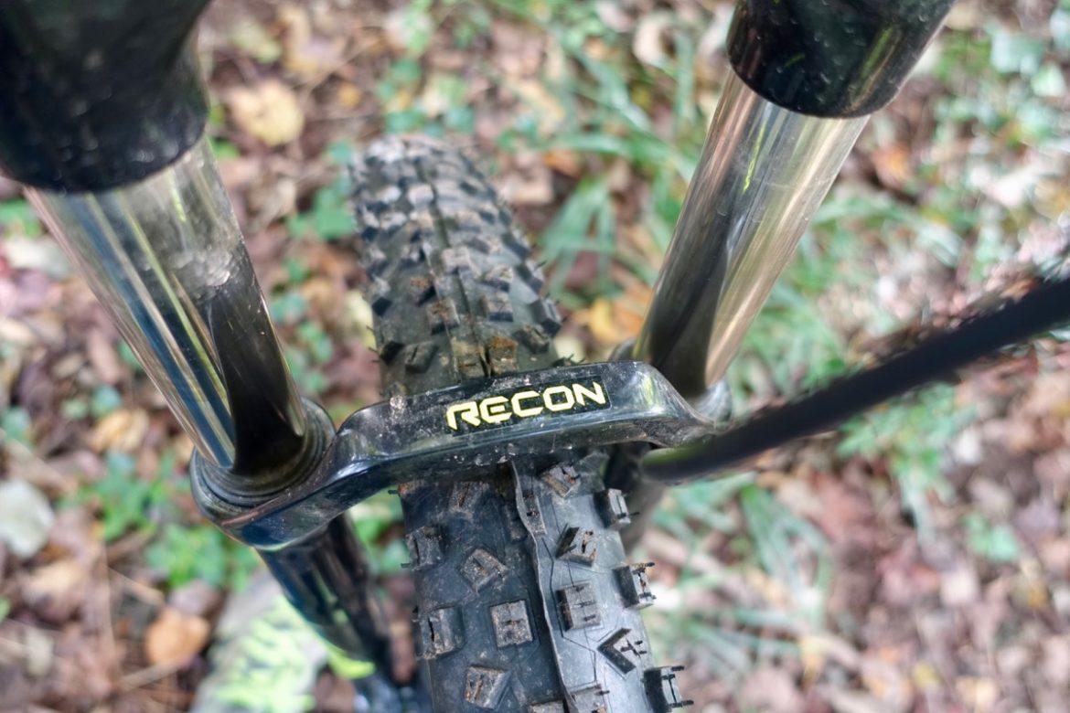 Marin San Quentin 2: Close-up view of a mountain bike's front fork and tire, featuring the brand name "RECON" prominently displayed. The background is a natural setting with fallen leaves and green foliage, indicating an outdoor environment. The tire shows rugged tread suitable for off-road terrain.