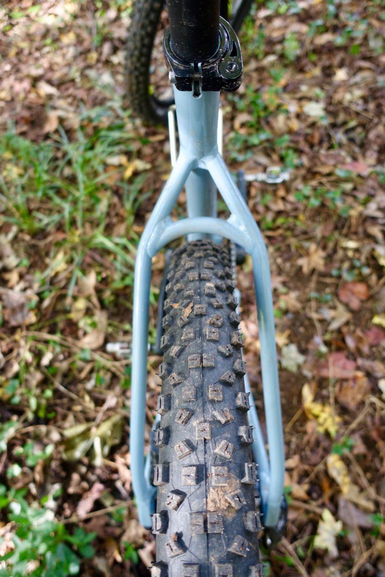 Marin San Quentin 2: Close-up view of a mountain bike's rear tire showing its textured, knobby surface, with a focus on the bike frame and the surrounding natural environment, including grass and fallen leaves.