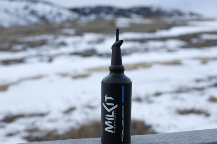 A black bottle with a nozzle, labeled "MILKit," sits on a ledge in a snowy landscape. The background features white snow covering the ground, with muted hills in the distance under an overcast sky.