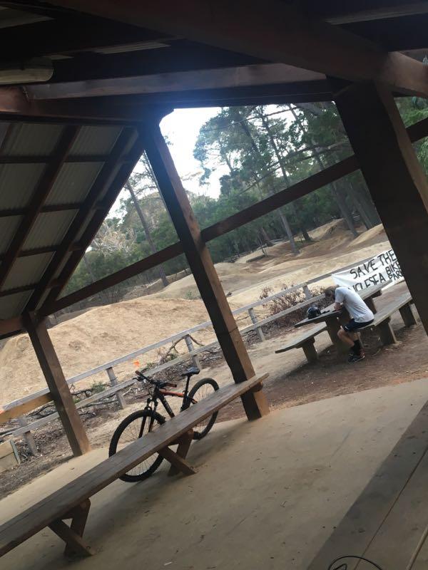 A covered pavilion at a bike park featuring a wooden table and bench. A mountain bike leans against the table, while a person sits at the table, looking down. In the background, dirt hills and trees are visible, along with a banner that reads "SAVE THE CHELA BIKES." The scene is set in an outdoor environment, emphasizing a community space for biking activities. Anglesea Mountain Bike Park mountain bike trail.