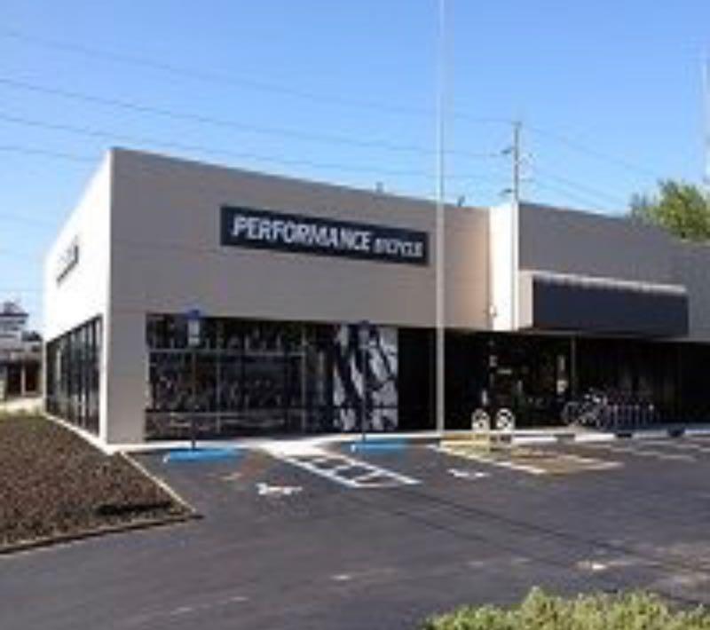 A modern bicycle shop with large windows, prominently displaying the sign "PERFORMANCE BICYCLE." The storefront features an open parking area with bicycle racks and clear blue skies above.