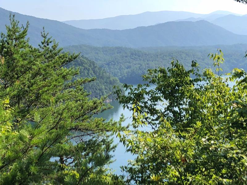 Scenic view of mountains and a lake, framed by lush green trees under a clear blue sky. The landscape shows rolling hills in the background, with water visible in the foreground, creating a serene natural setting. Tsali Recreation Area mountain bike trail.
