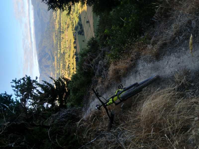 A mountain bike rests on a dirt trail surrounded by vegetation, overlooking a scenic valley with fields and mountains in the background. The image captures a rugged terrain under clear skies. Rameka Track mountain bike trail.