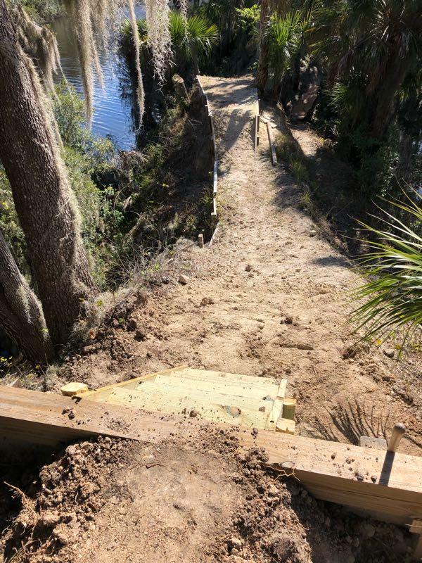 A sandy, recently constructed pathway leading down to a body of water, flanked by trees and vegetation. The pathway features wooden steps at the top, and the surrounding area shows disturbed earth, indicating ongoing landscaping or construction work. Loyce E. Harpe Park mountain bike trail.