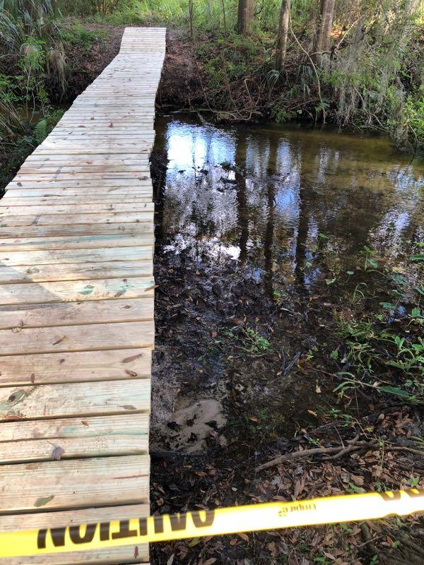 A wooden bridge leading over a small creek, surrounded by dense greenery and trees. A caution tape is visible in the foreground, indicating a restricted area. The water reflects the surrounding environment. Loyce E. Harpe Park mountain bike trail.