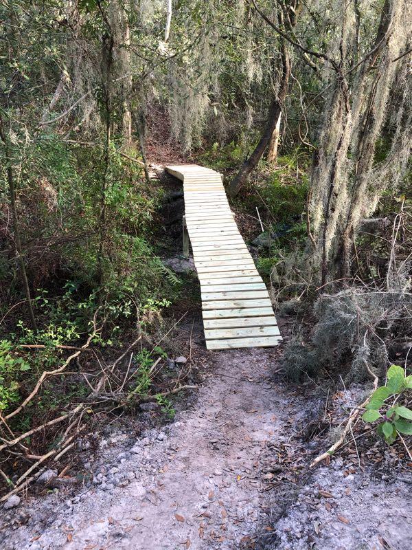 A wooden boardwalk extends through a lush, green forest, surrounded by trees and hanging Spanish moss. The path leads slightly upward from a dirt trail, providing access to the natural surroundings. Loyce E. Harpe Park mountain bike trail.