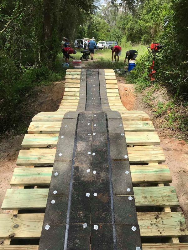 Construction of a wooden path or ramp in a wooded area. The structure is made of wooden planks arranged in a slightly curved shape, with a black surface in the center. Several people are working in the background, engaging in various tasks related to the construction. Tools and equipment are visible, with greenery surrounding the scene. Loyce E. Harpe Park mountain bike trail.