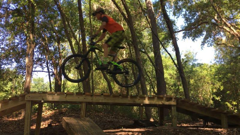 A person wearing an orange shirt and green patterned shorts is mid-jump on a mountain bike, soaring over a wooden bridge in a wooded area. The surrounding trees provide a lush background, highlighting an outdoor biking adventure. Loyce E. Harpe Park mountain bike trail.