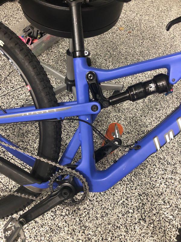 Close-up view of a blue mountain bike frame showcasing the rear suspension, drivetrain, and pedal assembly. The bike is positioned on a textured gray floor, highlighting its intricate components and design. Markham Park mountain bike trail.
