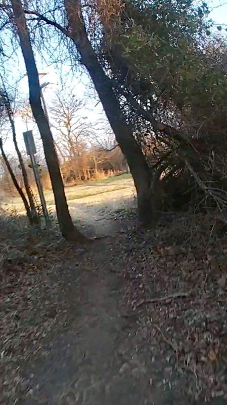 A narrow dirt path partially surrounded by trees and brush, leading to an open grassy area in the background. The scene is bathed in natural light, suggesting an early morning or late afternoon setting. A signpost is visible on the left side of the path. Chisenhall trails mountain bike trail.