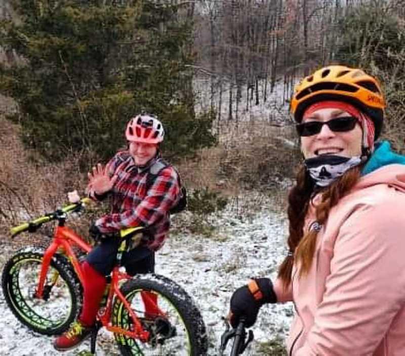 A man and a woman smiling while standing next to their fat bikes in a snowy, forested area. The man is wearing a red plaid shirt and a red helmet, waving at the camera. The woman is dressed in a pink hoodie, sunglasses, and a scarf, with long hair cascading down. Snow covers the ground, and trees are visible in the background. DTE Energy Foundation Trail mountain bike trail.