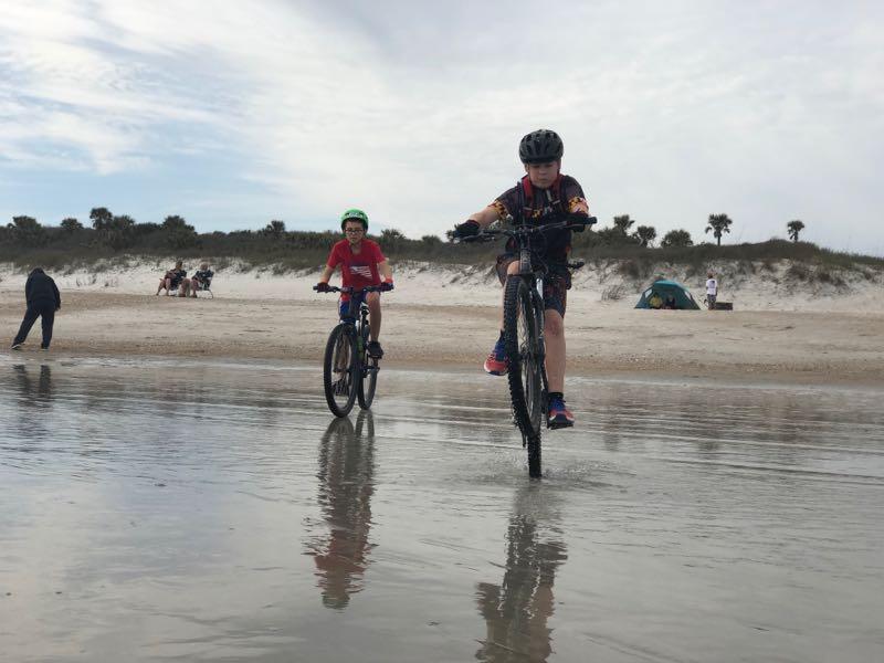 Two children riding bicycles along the shoreline at the beach, with one child lifting their front wheel in a playful maneuver. The sandy beach features small waves, and in the background, people are relaxing, along with a green tent and palm trees. The sky is partly cloudy, creating a serene atmosphere. Kathryn Abby Hanna Park mountain bike trail.