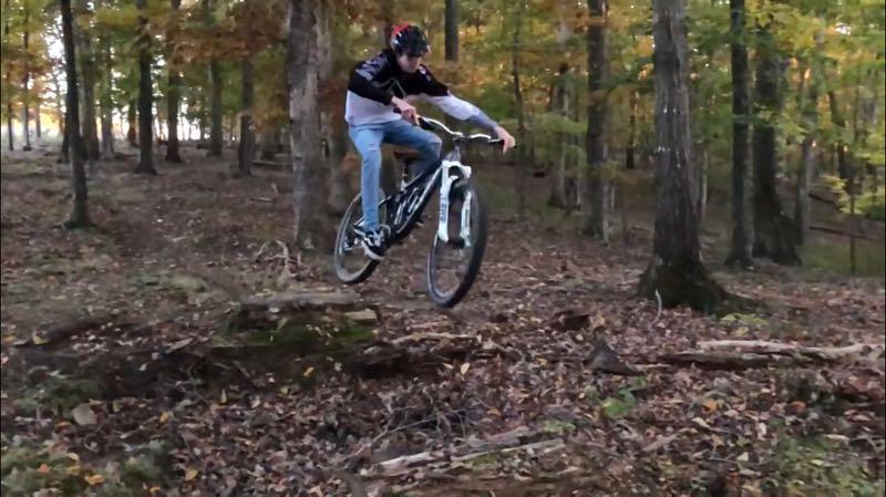 A person in a helmet jumps off a small wooden ramp while riding a mountain bike in a forested area with autumn foliage. Centerville Power Lines mountain bike trail.