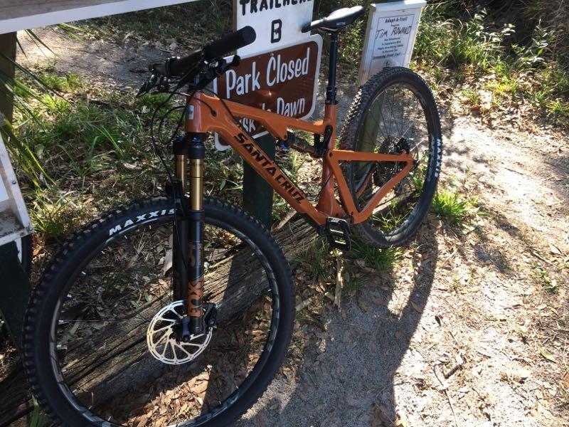 A mountain bike with a distinctive orange frame, parked next to a sign that reads "Park Closed Dawn to Dusk." The bike features front suspension and is positioned on a dirt path surrounded by greenery. Chuck Lennon Park mountain bike trail.