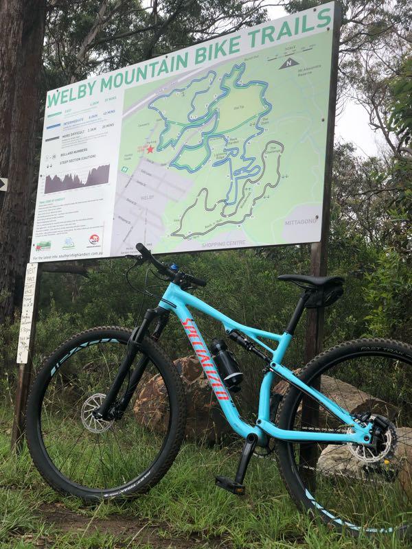 A blue mountain bike parked in front of a sign displaying the map and information for the Welby Mountain Bike Trails, surrounded by trees and natural scenery. Welby Cemetery mountain bike trail.