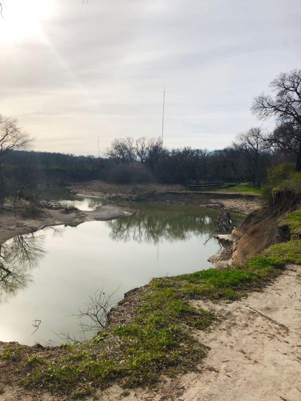 A tranquil landscape featuring a calm river surrounded by barren trees and grassy banks. The water reflects the overcast sky, while a distant radio tower is faintly visible in the background. The scene captures a peaceful natural setting, ideal for outdoor activities. Gateway Park mountain bike trail.