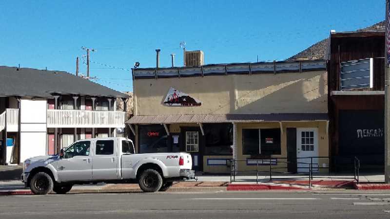 A silver pickup truck is parked in front of a single-story building with a yellow exterior and a corrugated metal awning. The building has multiple windows, and an "Open" sign is visible. There is a small ramp leading to the entrance, and two additional buildings are seen in the background, one of which is multi-story with balconies. The sky is clear and blue, indicating a sunny day.