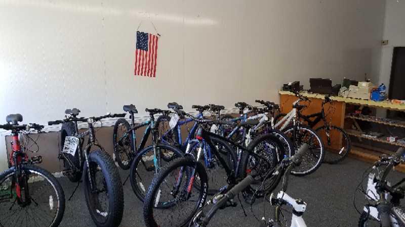 A view of a bike shop interior featuring several bicycles of various styles lined up on the floor. An American flag hangs on the wall in the background, and there is a counter with supplies visible. The floor is carpeted, and the atmosphere suggests a retail space focused on cycling products.