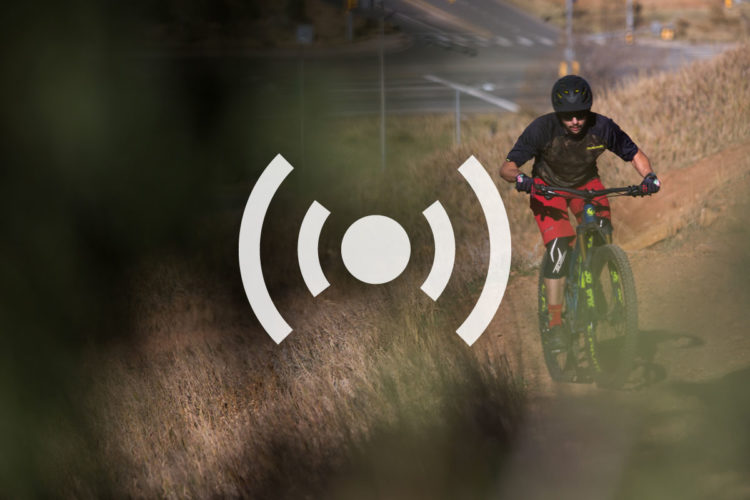 A mountain biker riding along a dirt trail surrounded by grass, with a blurred background featuring a road. An overlay graphic of a signal icon is positioned in the center.
