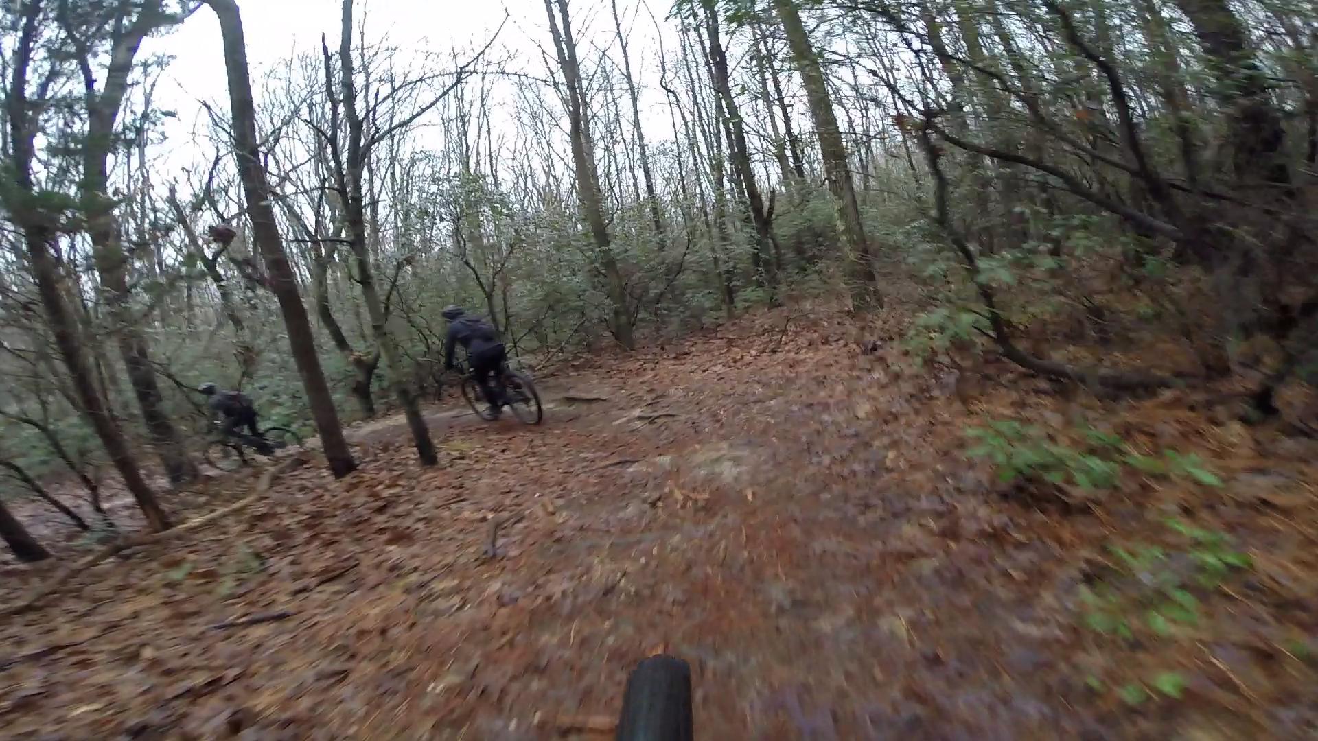 A mountain biker riding through a wooded trail covered in fallen leaves, with wet conditions and bare trees surrounding the path. The focus is on the bike and trail, conveying a sense of adventure and nature. Allaire State Park mountain bike trail.