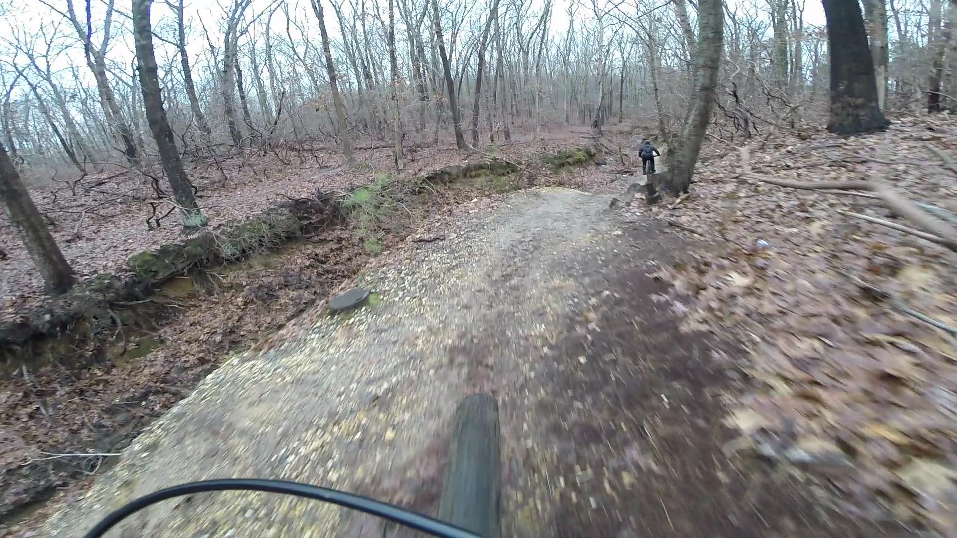 Mountain biking on a gravel trail surrounded by bare trees and fallen leaves in a wooded area. A cyclist is seen riding in the distance, with the bicycle tire in the foreground showing the uneven terrain. Allaire State Park mountain bike trail.