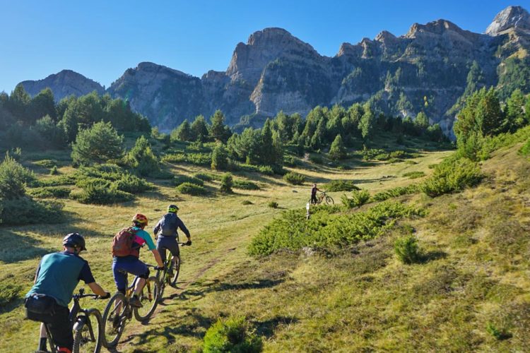 A group of four mountain bikers riding along a scenic dirt trail in a mountainous landscape. The setting features lush green vegetation and towering rocky mountains under a clear blue sky. One cyclist is ahead, while another is seen walking with their bike in a slightly elevated area surrounded by trees.