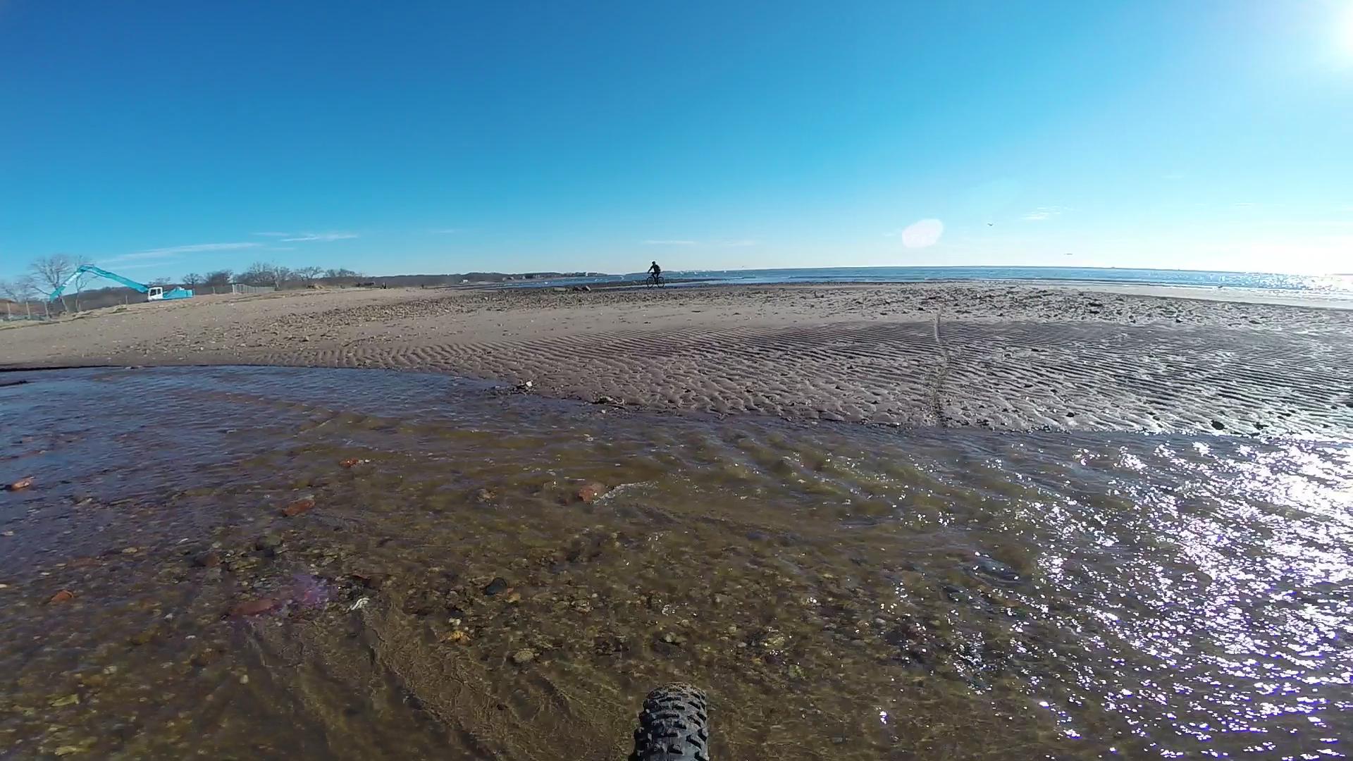 A scenic view of a beach with rippled sand and shallow water in the foreground. In the background, a cyclist rides along the shoreline, with a clear blue sky overhead and the ocean visible in the distance. A blue construction crane is positioned near the shore, indicating some ongoing activity in the area. Wolfes Pond park mountain bike trail.