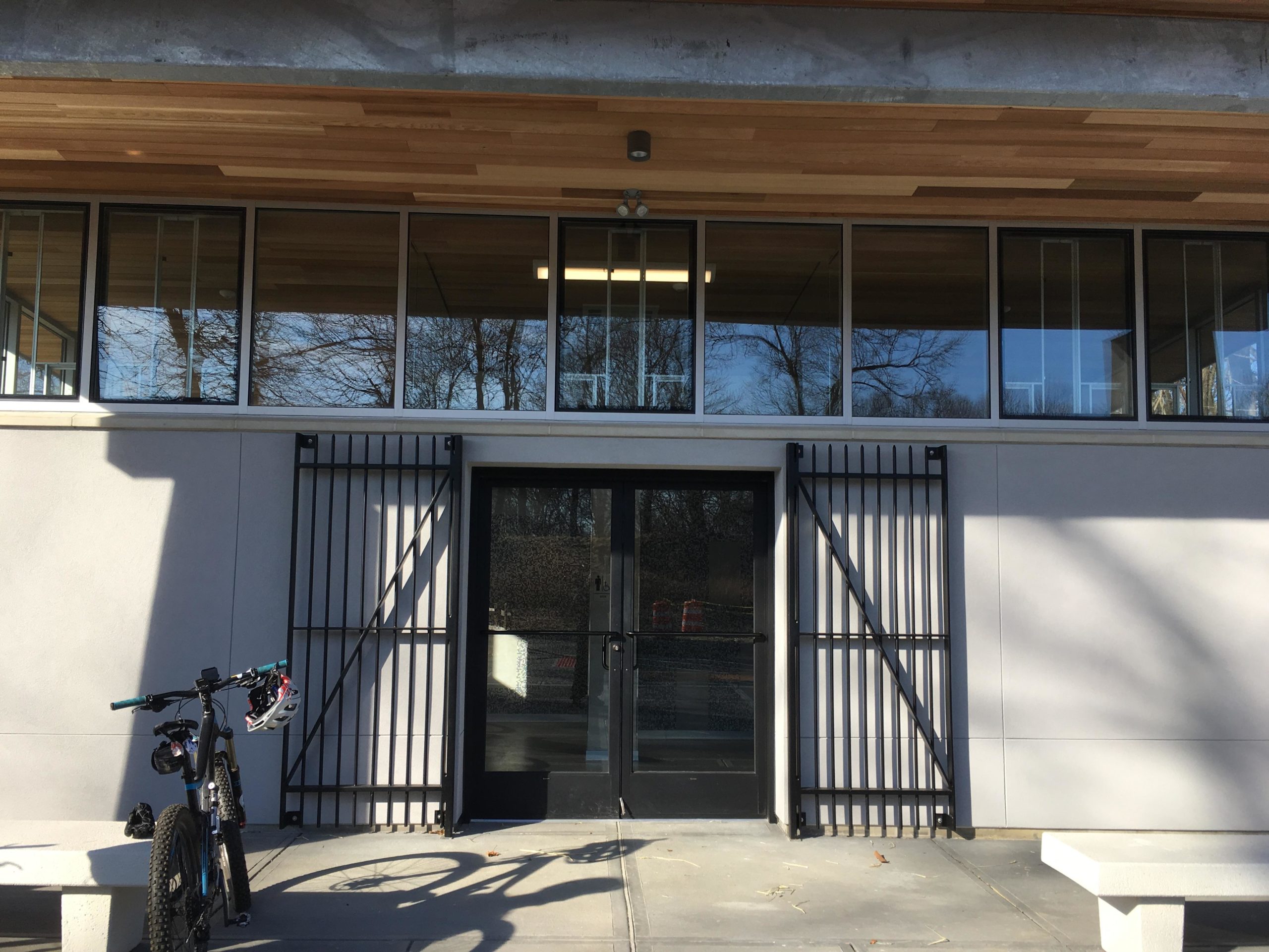 A modern building facade featuring large windows, double glass doors, and decorative metal grates on either side of the entrance. A mountain bike is parked in the foreground, casting a shadow on the concrete walkway. The building has a wooden ceiling above the entrance, with trees visible through the windows. Hartshorne Woods Park mountain bike trail.