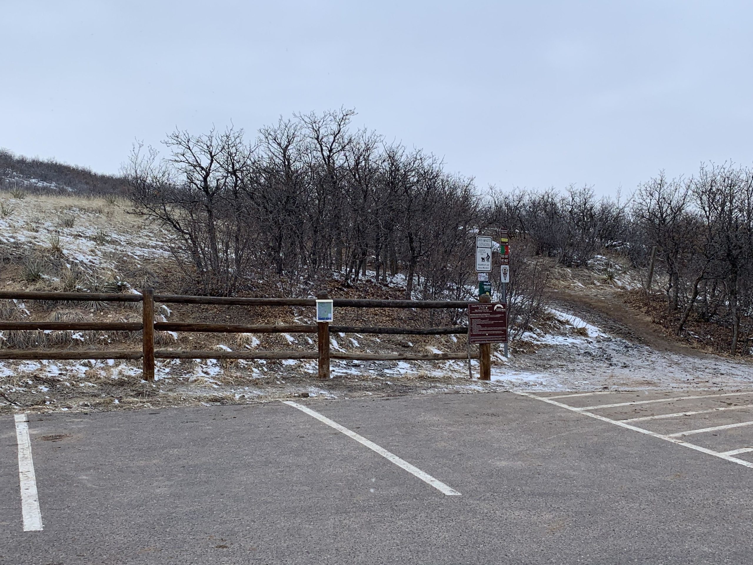 A parking area at a trailhead on a cloudy day, featuring a wooden fence and a sandy path leading into a wooded area. Snow can be seen on the ground, and there are several informational signs about the trail. The surrounding landscape is mostly barren, with sparse trees and dry grass. Ridgeline Open Space Trail mountain bike trail.