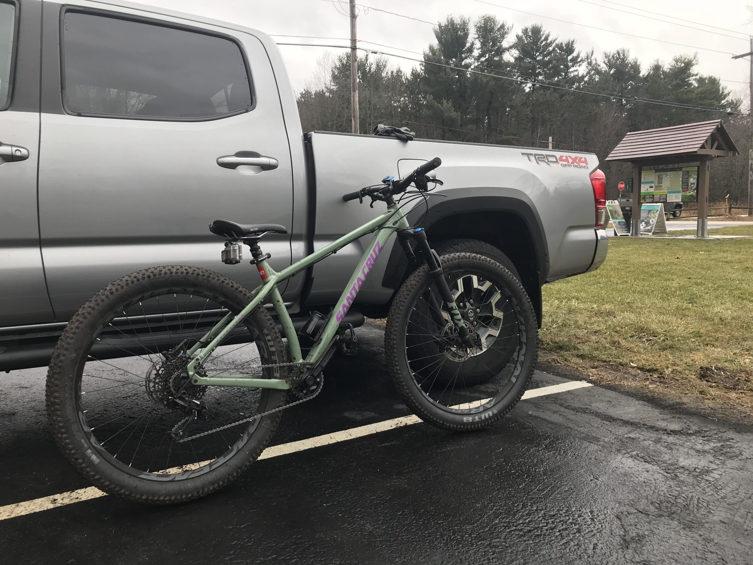 Santa Cruz Chameleon: A mountain bike is parked next to a silver pickup truck in a parking lot. The bike has a green frame with purple lettering and features thick tires suitable for off-road riding. In the background, there is a wooden information kiosk with signage, surrounded by grass and trees. The scene is overcast, suggesting a cloudy day.