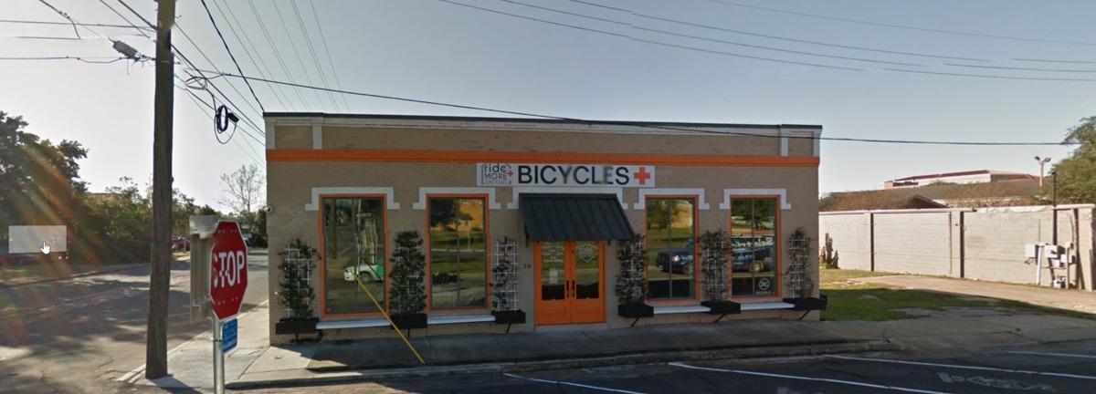 A brick storefront featuring large windows and an orange accent stripe, labeled "ride + MORE BICYCLES." The entrance has a black awning, and there are decorative plants in front. Nearby traffic signs and power lines are visible in a clear blue sky.